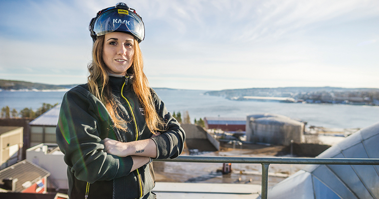 Kvinna i arbetskläder och skyddshjälm med glasögon. Fotad på en byggnad med kraftvärmeverket i Härnösand i bakgrunden.